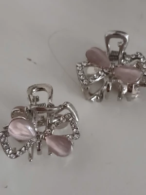 Söta hårklämmor med rosett och stenar - Två silvriga hårklämmor formade som rosetter med ljusrosa pärlemorskimrande detaljer och glittrande stenar. Perfekta för att piffa till frisyren med en söt och feminin touch. Metallbas med dekorativa inslag som ger ett lyxigt intryck.