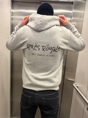 Grå hoodie Après Royaleä Nolt - Säljer en ljusgrå hoodie från Après Royale med tryck på ryggen där det står 'APRÈS Royale' och 'NOLT NORDIC ALPINE'. Hoodien har en stor huva och är gjord i mjukt material, perfekt för chill dagar. Klassisk passform och ribbade muddar.