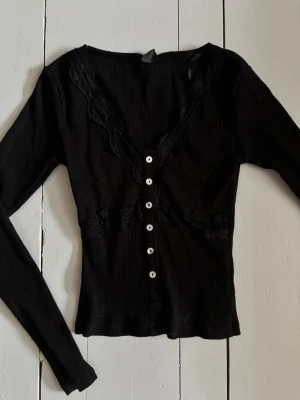 Svart ribbad topp med spets - Snygg svart ribbad topp med långa ärmar, v-ringning och spetsdetaljer längs halsringningen. Toppen har söta vita knappar framtill och sitter tight. Perfekt att styla med ett par jeans!
