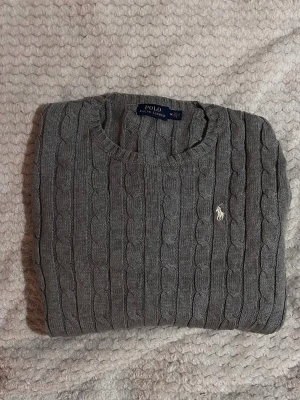 Grå kabelstickad tröja Polo Ralph Lauren - Kabelstickad grå tröja från Polo Ralph Lauren med rund halsringning och lång ärm. Tröjan har det klassiska broderade logotypen på bröstet och ribbade muddar vid ärmslut och nederkant. Perfekt för lager på lager och stilren look.