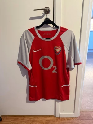 Arsenal matchtröja Nike röd/grå - Säljer en klassisk Arsenal matchtröja från Nike i rött och grått med klubbmärke och O2-logga på bröstet. Tröjan har korta ärmar, rund hals och är tillverkad i ett lätt, ventilerande material perfekt för fotboll eller träning.