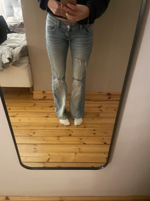 Ljusblå bootcut jeans med slitningar - Säljer ett par ljusblå bootcut jeans med slitningar på låret och knäet. Jeansen har låg midja, klassiska fem fickor och snygga detaljer på bakfickorna. Perfekta för en avslappnad och trendig look.