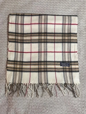 Rutig ullhalsduk från Burberrys - Klassisk halsduk i 100% lammull från Burberrys med ikoniskt rutmönster i beige, svart, vitt och röda detaljer. Halsduken har fransar i ändarna och en vävd etikett. Perfekt accessoar för att lyfta din outfit med en tidlös brittisk stil.