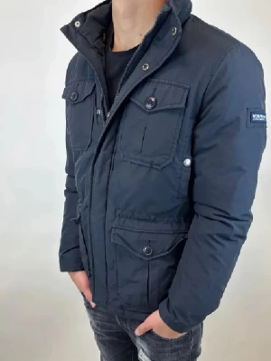 Mörkblå vindjacka från Woolrich - Snygg mörkblå vindjacka från Woolrich med fyra stora fickor framtill och dragkedja samt knappar. Jackan har hög krage och patch med logga på ärmen. Perfekt för dig som vill ha en stilren och funktionell jacka till vardags.