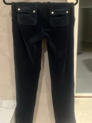 Svarta velourbyxor Juicy Couture - Mörkblå mjukisbyxor från Juicy Couture i klassisk velour med raka ben och två bakfickor med lock och silverknappar. Byxorna har dragsko i midjan och är perfekta för en chill och avslappnad stil. Lite smutsiga som syns på bild men de försvinner i tvätt