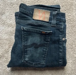 Nudie jeans - Säljer ett par svarta jeans från Nudie Jeans med klassisk design och snygga detaljer på bakfickorna. Storleken på jeansen är W30 L32. Jeansen har en slim passform och är tillverkade i mjukt denimtyg med lätt tvättad utseende. Priset kan diskuteras