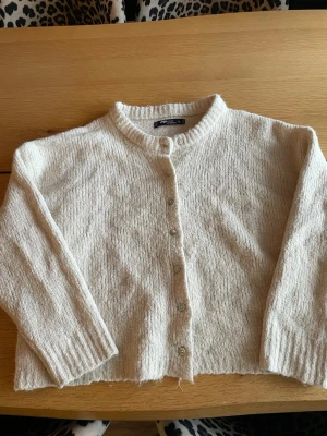 Beige stickad kofta  - Mysig beige stickad kofta med rund halsringning och knappar framtill. Koftan har lång ärm och ribbade muddar vid ärmslut och nederkant. Perfekt att slänga över en t-shirt eller linne för en chill look.