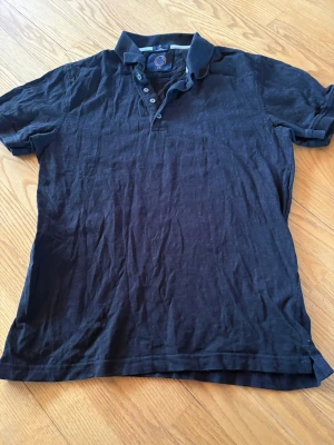 Svart pikétröja från Human Scales - Klassisk svart pikétröja från Human Scales med krage och knappar framtill. Tröjan har korta ärmar och är tillverkad i mjukt bomullstyg. Perfekt till jeans eller shorts för en avslappnad look.