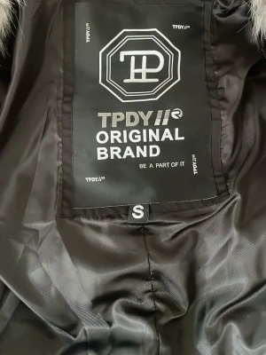 Svart parkas från TPDY  - Snygg svart parkas från TPDY Original Brand med stora grå fuskpälsdetaljer vid huvan. Jackan har en klassisk parkasdesign och är fodrad för extra värme. Perfekt för kalla dagar och ger en cool streetstyle-look.