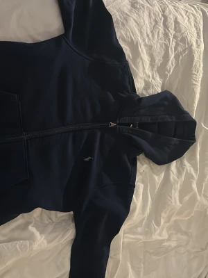 Mörkblå hoodie från Polo Ralph Lauren - Snygg mörkblå hoodie från Polo Ralph Lauren med dragkedja framtill och klassisk huva. Ikoniska loggan broderad på bröstet. Tillverkad i mjukt bomullsmaterial och har fickor på sidorna. Perfekt för en chill och stilren look.