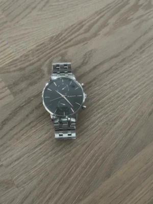 GANT herrklocka med svart urtavla - Stilren herrklocka från GANT med svart urtavla och silverfärgat armband i metall. Klockan har kronograf-funktion och tydliga markeringar på urtavlan. Perfekt accessoar för dig som gillar klassisk och modern stil.