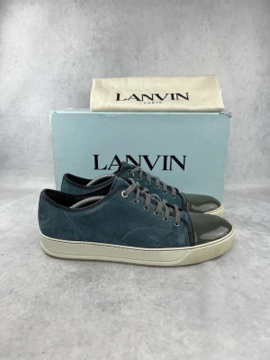 Blå Lanvin sneakers med lackad tå - Snygga sneakers från Lanvin i blå mocka med grå lackad tå och vita sulor. Skorna har grå skosnören och en klassisk låg profil. Perfekta för dig som gillar stilrena och exklusiva detaljer. Kommer med originalkartong och dustbag.