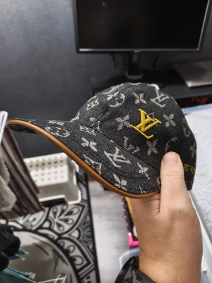 Louis Vuitton monogram keps svart - Säljer en svart keps från Louis Vuitton med klassiskt monogrammönster i grått och en gul LV-logga framtill. Kepsen har böjd skärm med brun kant och är tillverkad i ett denimliknande material. Perfekt för dig som vill ha en exklusiv och trendig accessoar.