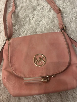 Rosa axelväska från Michael Kors - Snygg rosa axelväska från Michael Kors i mjukt konstläder. Väskan har justerbar axelrem, guldfärgade detaljer och en dekorativ tofs. Framsidan pryds av MK-loggan i metall. Perfekt storlek för det viktigaste och stängs med dragkedja under locket. Den är även väldigt stor och rymmlig med massor av olika fack🤍