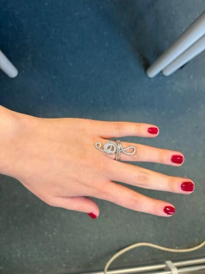 Silvrig ring med G-klav design - Unik ring i silverfärgad metall formad som en G-klav, perfekt för dig som älskar musik och vill sticka ut. Ringen har en cool, handgjord känsla med snirkliga detaljer och passar både till vardagslooken och när du vill addera något extra till din stil.