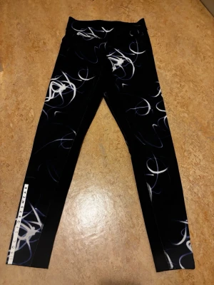 Svarta leggings från SOC Sportswear - Snygga svarta leggings från SOC Sportswear med vitt och blått abstrakt mönster. Tight passform och elastiskt material som passar perfekt till träning eller chill. Logga längs benet för en sportig vibe.