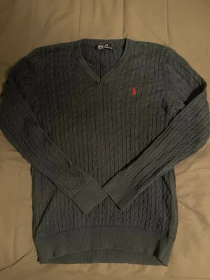 Mörkgrå kabelstickad tröja Polo Ralph Lauren - Snygg mörkgrå kabelstickad tröja från Polo Ralph Lauren med V-ringning och klassisk röd logga broderad på bröstet. Tröjan har långa ärmar och ribbade muddar vid ärmslut och nederkant. Perfekt för dig som gillar stilrena och tidlösa plagg.
