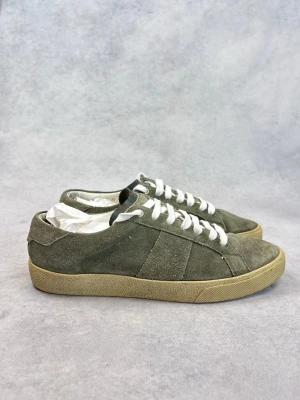 Olivgröna Saint Laurent mockasneakers med vita snören - Stilrena sneakers i olivgrön mocka med vita skosnören och beige gummisula. Klassisk låg modell med enkel design som passar till många outfits. Perfekta för dig som gillar en clean och avslappnad stil.