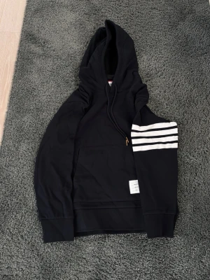 Svart hoodie från Thom Browne - Snygg svart hoodie från Thom Browne med klassiska vita ränder på ena ärmen och en liten etikett vid nederkanten. Hoodien har huva med dragsko och en stor magficka. Tillverkad i Japan och har en exklusiv känsla. Perfekt för dig som gillar streetwear och stilrena detaljer.