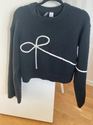 Svart cropped stickad tröja Gina Tricot - Säljer en svart cropped stickad tröja från Gina Tricot med ett vitt dekorativt band i form av en rosett framtill. Tröjan har rund halsringning, långa ärmar och ribbade muddar. Perfekt för dig som gillar trendiga detaljer och en avslappnad look.