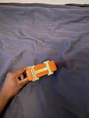 Orange H bälte med guld H spänne  - Snyggt brunt skärp med ett stort, guldfärgat H-format spänne. Skärpet har en stilren design och är tillverkat i skinn med vita sömmar längs kanterna. Perfekt accessoar för att lyfta din outfit.