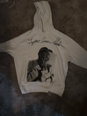 Vit hoodie med Tupac-tryck - Säljer en vit hoodie med stor svartvit bild av Tupac Shakur på framsidan och hans signatur ovanför. Hoodien har huva och ribbade muddar. Perfekt för dig som gillar streetwear och vill ha en unik look.