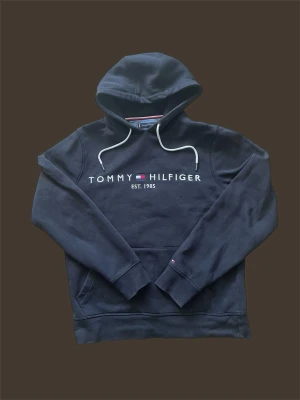 Svart hoodie från Tommy Hilfiger - Snygg svart hoodie från Tommy Hilfiger med vit logga och text 'EST. 1985' på bröstet. Klassisk huva med vita dragsnören och magficka framtill. Tillverkad i mjukt bomullsmaterial, perfekt för en avslappnad stil.