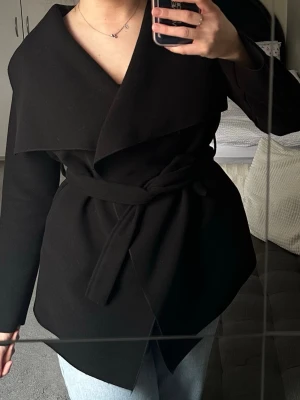 Svart omlottkappa med knytskärp - Stilren svart kappa i omlottmodell med brett slag och knytskärp i midjan. Kappan har en mjuk, slät yta och rak passform som ger en modern look. 