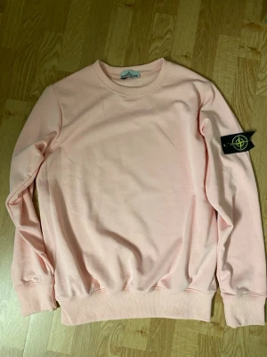 Ljusrosa sweatshirt från Stone Island - Ljusrosa sweatshirt från Stone Island med klassisk rund hals och ribbade muddar. Tröjan har den ikoniska svarta patchen med gul och grön kompasslogga på vänster ärm. Tillverkad i mjuk bomull för en skön och avslappnad look.