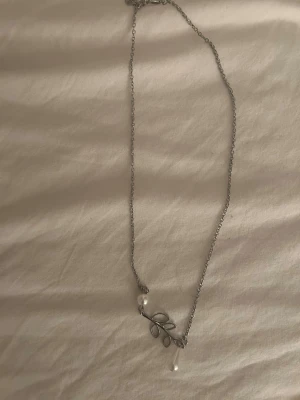 Silverfärgat halsband med pärldetaljer - Snyggt halsband i silverfärg med tunn kedja och ett hängsmycke format som en gren med blad. Två små vita pärlor sitter fast i ändarna på hänget och ger en elegant touch. Perfekt accessoar för att lyfta din outfit.