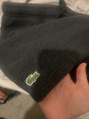 Mörkgrå shorts från Lacoste - Säljer ett par mörkgrå mjukisbyxor från Lacoste med regular fit. Byxorna har resår i midjan med dragsko och den klassiska gröna krokodilloggan broderad vid benet. Insidan är mjuk och fleecefodrad för extra komfort.