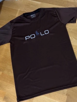 Brun POLO t-shirt - Snygg brun t-shirt från POLO med stort tryck på bröstet och klassisk logga. T-shirten har rund hals och är kortärmad, tillverkad i dri fit. Perfekt för en avslappnad och sportig stil. Storlek: M-L men passar mest M