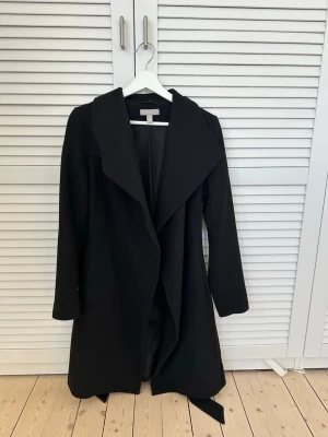 Svart kappa med skärp  - Stilren svart kappa med bred krage och knytskärp i midjan. Materialet är en ullblandning och kappan passar därmed perfekt till hösten/vintern. Längden slutar en bit nedanför knäna.