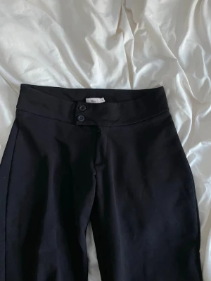 Svarta kostymbyxor från Nelly - Stilrena svarta kostymbyxor från Nelly med hög midja och raka ben. Byxorna har två knappar i midjan och är sydda i ett mjukt, vävt material som ger en snygg passform. Perfekta för en clean och trendig look.