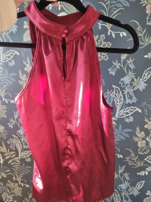Vinröd ärmlös halterneck-topp - Snygg vinröd halterneck-topp i glansigt satinliknande material. Toppen är ärmlös med hög krage och dekorativa knappar framtill. Perfekt för dig som vill ha en elegant och trendig look.