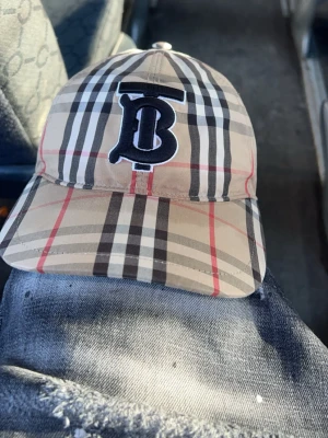 Beige Burberry keps med rutmönster - Säljer en klassisk Burberry keps i beige med det ikoniska rutmönstret i svart, vitt och rött. Framtill sitter ett stort svart broderat TB-monogram. Kepsen har böjd skärm och justerbar baksida. Perfekt för dig som vill ha en stilren och trendig accessoar.