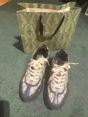 G U C C I sneakers med monogram och blå detaljer - Säljer ett par snygga G U C C I sneakers med klassiskt beige monogrammönster och mörkblå detaljer. Skorna har vita snören och tjock sula för en trendig look. Perfekta för dig som vill sticka ut med exklusiva och stilrena sneakers.