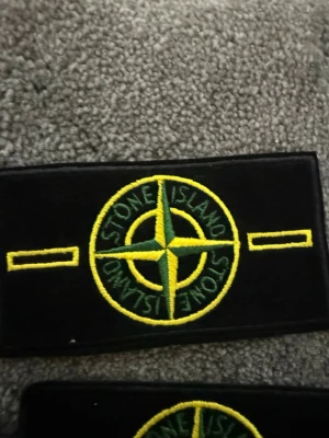 130kr/styck kan ävensänka liteStone Island tygmärke svart/gul - Säljer ett svart tygmärke från Stone Island med den ikoniska kompassen broderad i gult och grönt. Märket är rektangulärt och har texten 'Stone Island' runt kompassen. Perfekt att sy eller fästa på jackor, väskor eller kepsar.