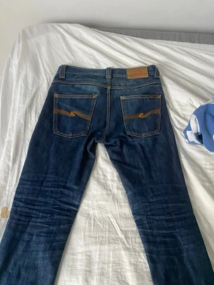 Mörkblå jeans från Nudie Jeans - Säljer ett par mörkblå jeans från Nudie Jeans med klassisk orange söm och vågmönster på bakfickorna. Jeansen har rak passform och är tillverkade i slitstark denim. Perfekta för dig som gillar en tidlös och snygg look.