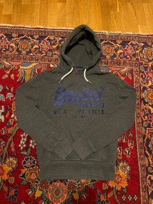 Mörkgrå Superdry hoodie med tryck - Mörkgrå hoodie från Superdry med blått tryck på bröstet och textdetaljer. Klassisk huva med vita dragsnören och känguruficka framtill. Mjuk bomullsblandning och ribbade muddar vid ärmslut och nederkant. Perfekt för en avslappnad streetwear-look.