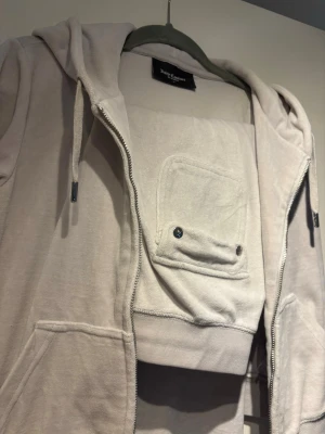 Grå set från Juicy Couture - Säljer ett grått sett från Juicy Couture med dragkedja, huva med snören och fickor framtill. Hoodien har en unik detalj med en ficka på bröstet och JC-logga på dragkedjan. Tillverkad i mjukt bomullsmaterial för en avslappnad och trendig look. Kofta i st S och byxor i M, pris kan diskuteras vid snabb affär och kan skickas eller mötas upp i Stockholm 