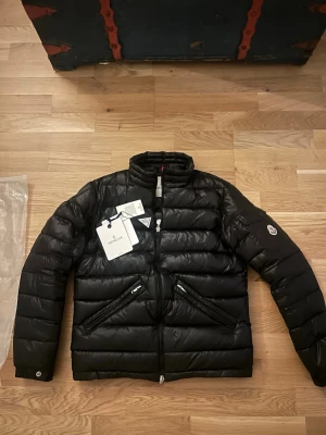 Svart dunjacka från Moncler - Snygg svart dunjacka från Moncler med hög krage och två dragkedjeförsedda fickor framtill. Jackan har quiltad design och klassisk Moncler-logga på ärmen. Perfekt för kalla vinterdagar och tillverkad i glansigt material.