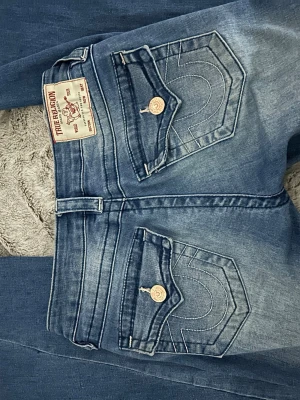Blå True Religion jeans - Säljer ett par blå jeans från True Religion med ikoniska ficklock bak och vita kontrastsömmar. W24 och lagom långa för mig som är 168. Köpt för ca 1300💓Väldigt bra skick men små revor på låren, de sista bilden