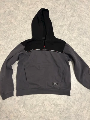Grå och svart hoodie från EA7 - Snygg hoodie från EA7 i grått och svart med halv dragkedja och huva. Dekorativt EA7-band över bröstet och logga nertill. Tillverkad i mjukt sweatshirtmaterial, perfekt för en avslappnad streetwear-look.