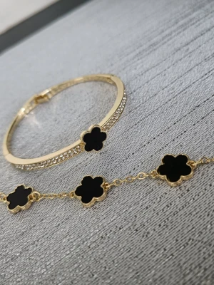 Guldarmband med svarta blommor - Snyggt armband i guldfärgad metall med svarta fyrklöverformade detaljer. Ena armbandet har en kedja med flera blommor, det andra är ett stelt armband med en blomma och gnistrande stenar längs kanten. Perfekt accessoar för att lyfta din stil.