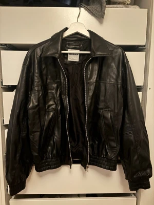 Svart skinnjacka från Bershka - Snygg svart skinnjacka från Bershka med klassisk krage, dragkedja framtill och två snedställda fickor. Jackan har resår vid midja och ärmslut för en schysst passform. 