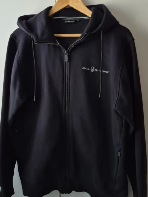 Sail Racing hoodie med dragkedja - Säljer en svart hoodie med dragkedja från Sail Racing med dragkedja framtill och justerbar huva med snören. Diskret logga på bröstet. Hoodien har fickor på sidorna och är tillverkad i mjukt bomullsmaterial. Perfekt för en avslappnad och sportig stil.
