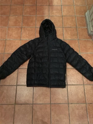 Svart dunjacka med huva från Peak Performance - Säljer en svart dunjacka från Peak Performance med huva och dragkedja framtill. Jackan har en quiltad design och två sidofickor med dragkedja. Perfekt för kalla vinterdagar och riktigt skön att bära.