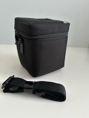 Sigma objektivväska / kameraväska  - Sigma objektivväska i svart tyg med dragkedja runt ovankanten. Väskan har vadderad insida med löstagbara innerskydd för att ge extra skydd åt objektivet. Utsidan är i slitstark textil med fästen för medföljande justerbar axelrem. Passar för förvaring och transport av kameraobjektiv eller annan liknande fotoutrustning. Helt NYTT!  Hämtas i Stockholm eller skickas mot porto. 