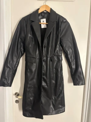 Svart lång kappa i fuskläder H&M - Stilren svart lång kappa från H&M i glansigt fuskläder. Klassisk krage, knäppning framtill med tre knappar och raka ärmar. Snygg och minimalistisk design som ger en edgy look. Perfekt för dig som vill ha en trendig och tidlös jacka.